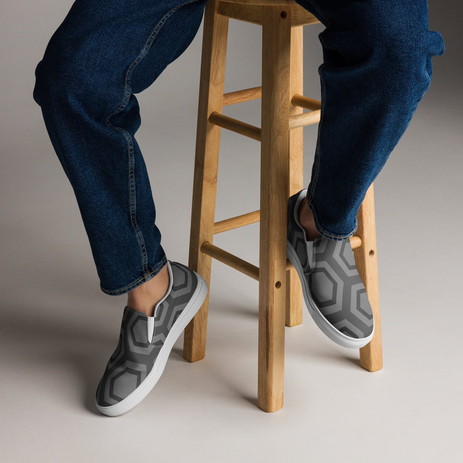 Person wearing trendy unisex clothing online with dark jeans and stylish gray slip-on shoes while sitting on wooden stool